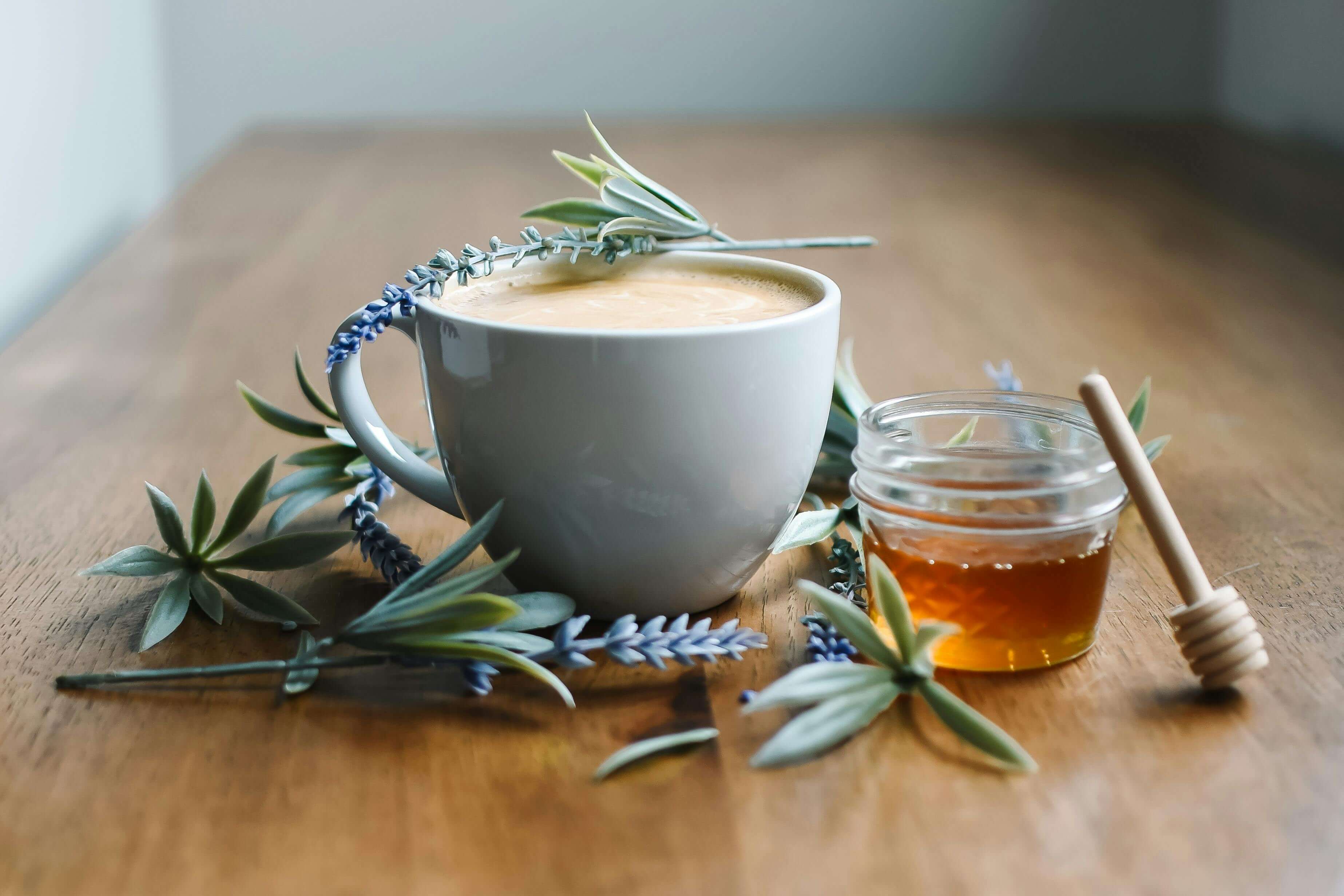 A white mug of coffee beside a small jar of honey with a dipper, surrounded by sprigs of lavender on a wooden table create a cozy, rustic feel.
